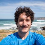A cheerful man with curly hair enjoys a sunny day by the ocean in Portugal.