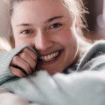 Portrait of cheerful young brunette putting head on arms folded on bed and looking at camera with open smile