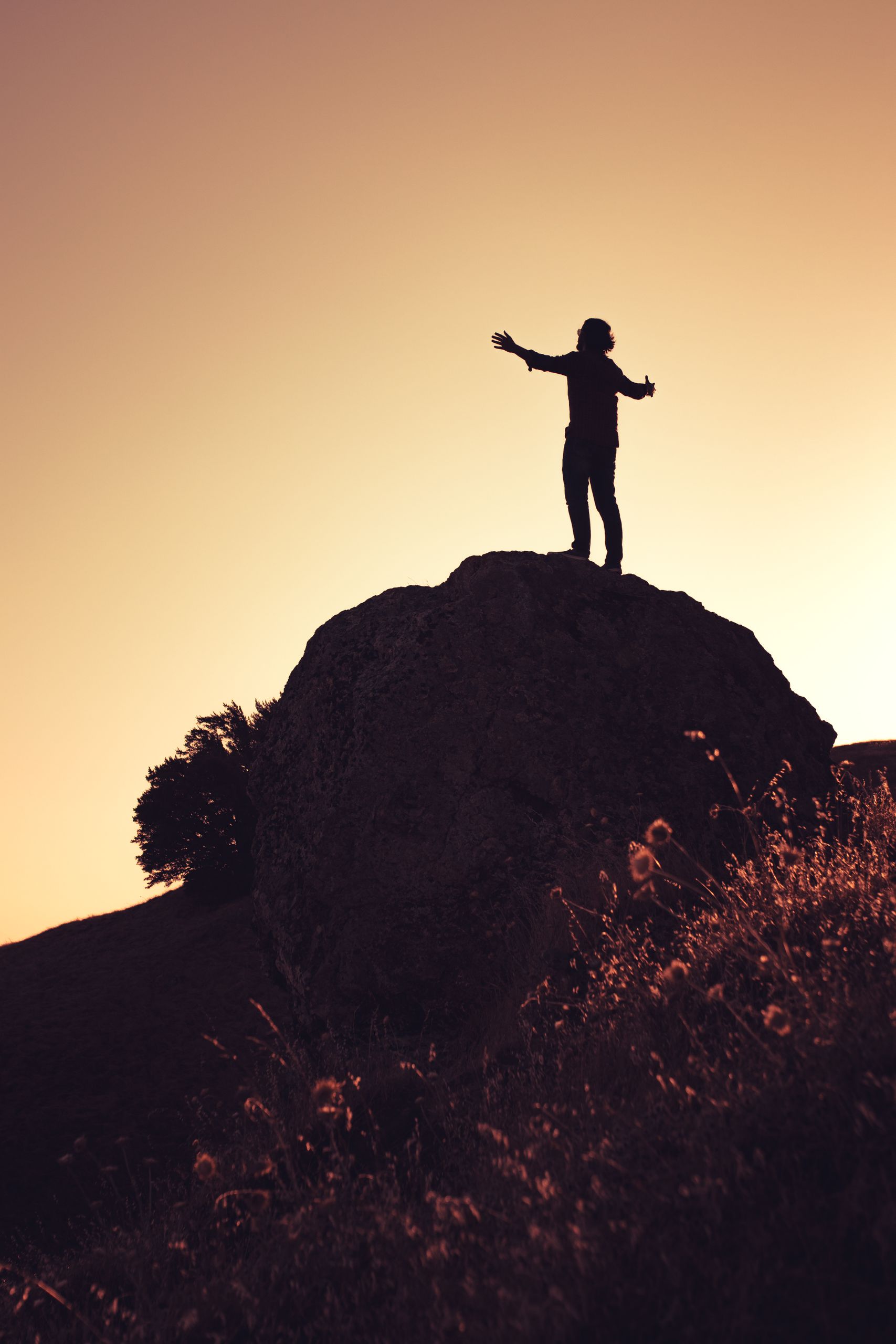Silhouette of an individual with arms outstretched atop a rock against a yellow sunset sky in California.